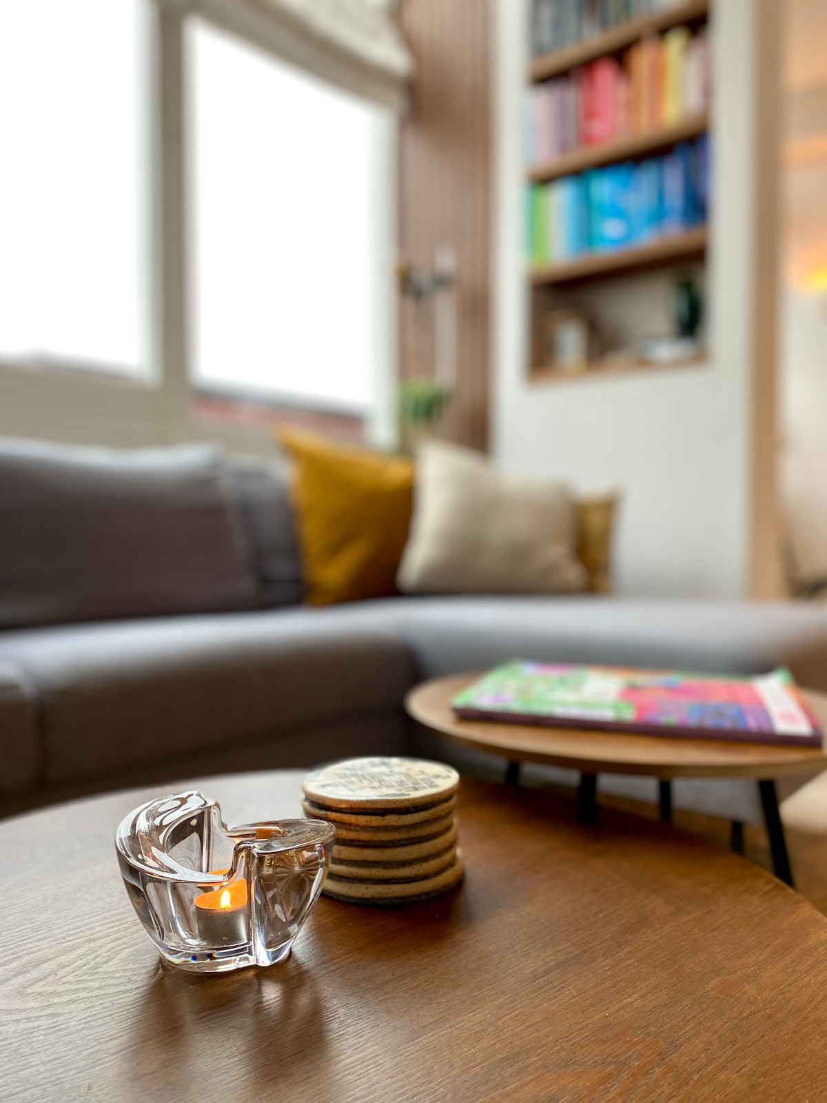 A cozy living space is seen with a comfortable gray sofa adorned with soft cushions. A wooden coffee table holds a decorative candle holder and a stack of coasters. In the background, a shelf displays an assortment of colorful books, creating an inviting atmosphere.