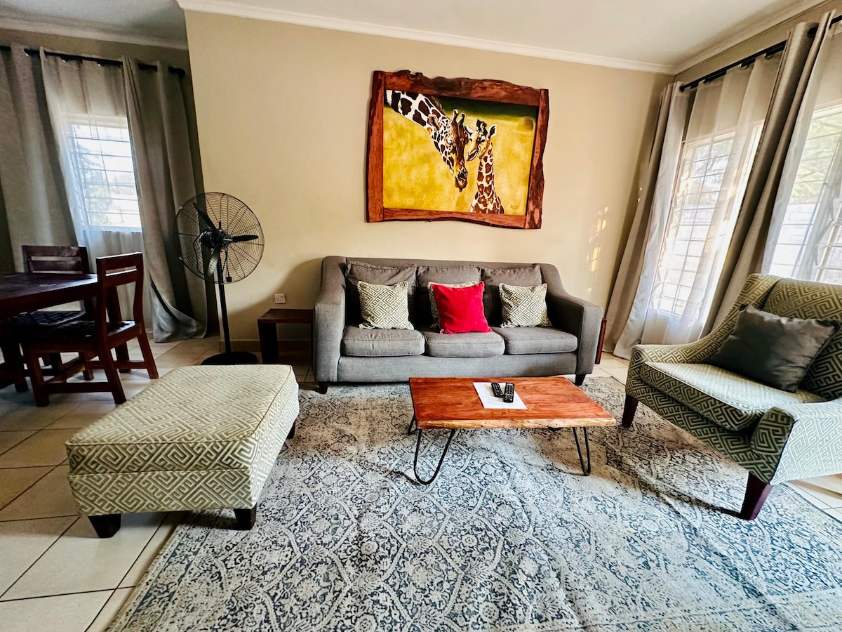 A comfortable lounge area features a neutral-toned sofa with a red accent pillow and two armchairs positioned around a wooden coffee table. A large piece of artwork depicting giraffes adorns the wall, complemented by light streaming through sheer curtains.