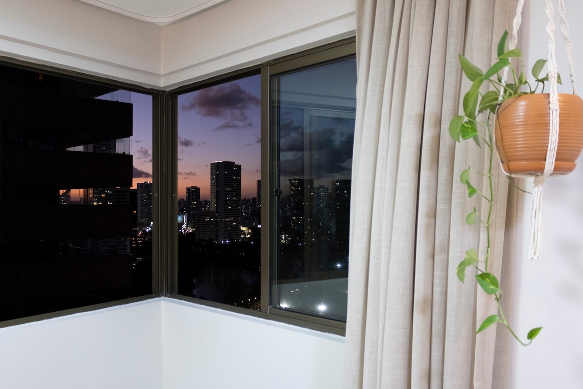 The corner of a room features large windows displaying a city skyline at dusk, with hues of orange and purple in the sky. A hanging plant adds a touch of greenery, while light fabric curtains softly frame the view.