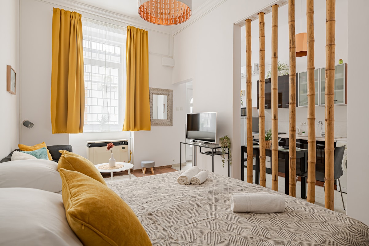 A spacious studio apartment features a comfortable double bed adorned with decorative pillows. Bright yellow curtains frame a large window, and a TV is positioned across from the bed. A kitchenette is visible in the background, separated by bamboo elements, adding warmth to the space.