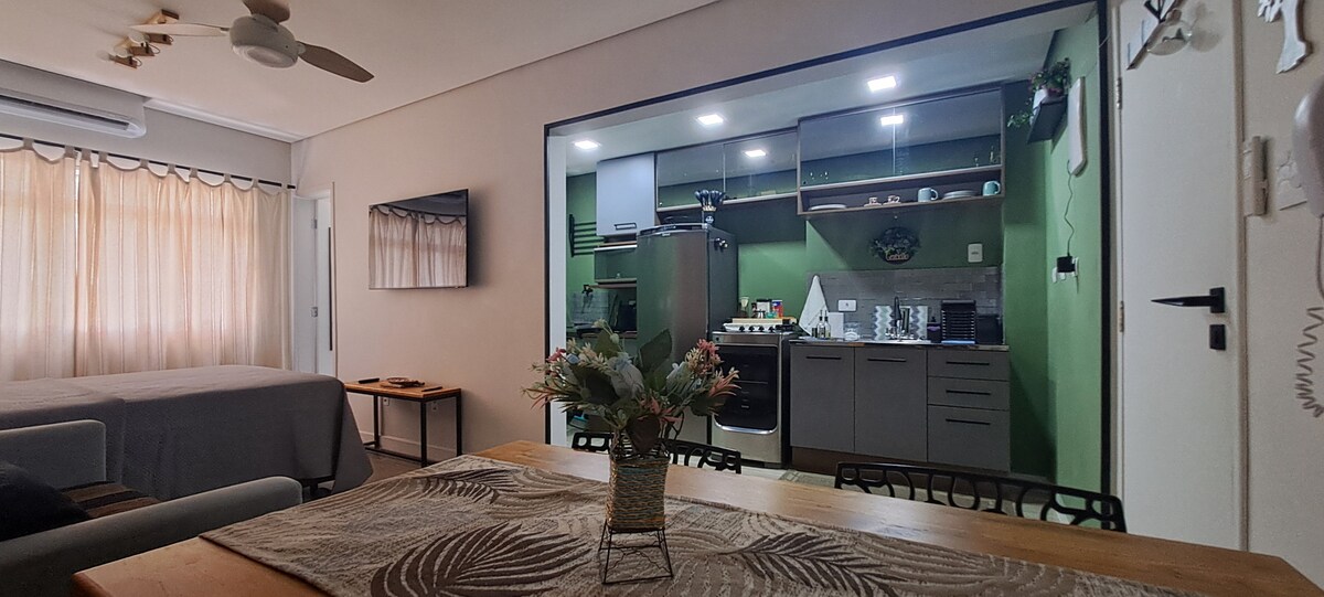A modern living area is connected to a stylish kitchen, featuring light green walls and sleek cabinets. A dining table is set with decorative plants, while a television is mounted on the wall. Natural light filters in through curtains, creating a bright and inviting space.