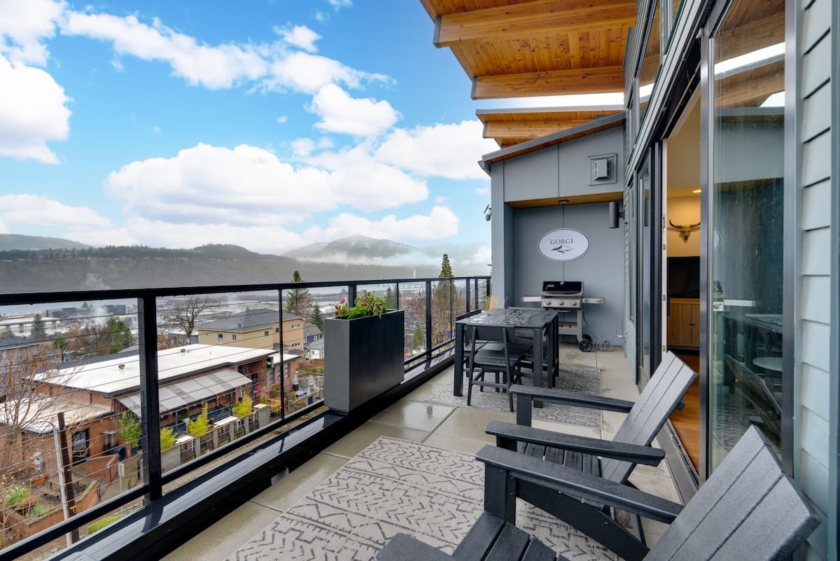 An outdoor deck offers a scenic view of the surrounding hills and the Columbia River. Black wooden seating is arranged on the deck, accompanied by potted plants. A gas grill is positioned against a wall, complementing the spacious area.