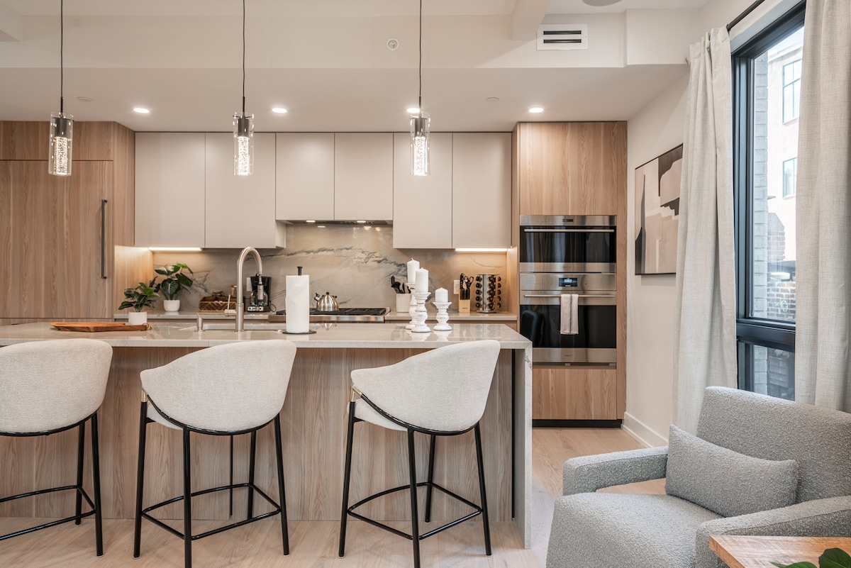 The modern kitchen features light wood cabinetry and a sleek countertop with three elegant bar stools. High-end appliances, including an oven and refrigerator, are integrated into the cabinetry. Large windows fill the space with natural light, enhancing the inviting atmosphere.
