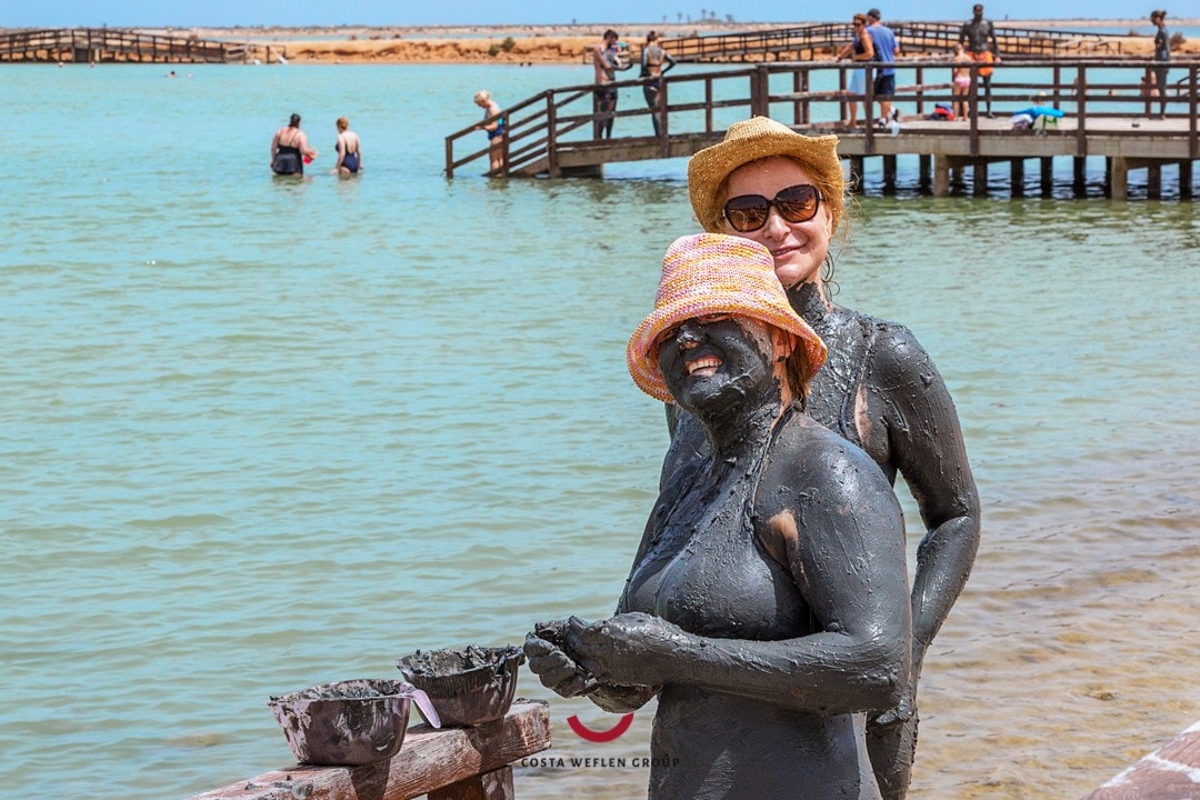 Two individuals are seen at a mud bath area, with mud applied to their skin. A wooden pier extends into the turquoise water, where people can be seen enjoying the surroundings. Sun hats are worn for sun protection, and the atmosphere appears lively.