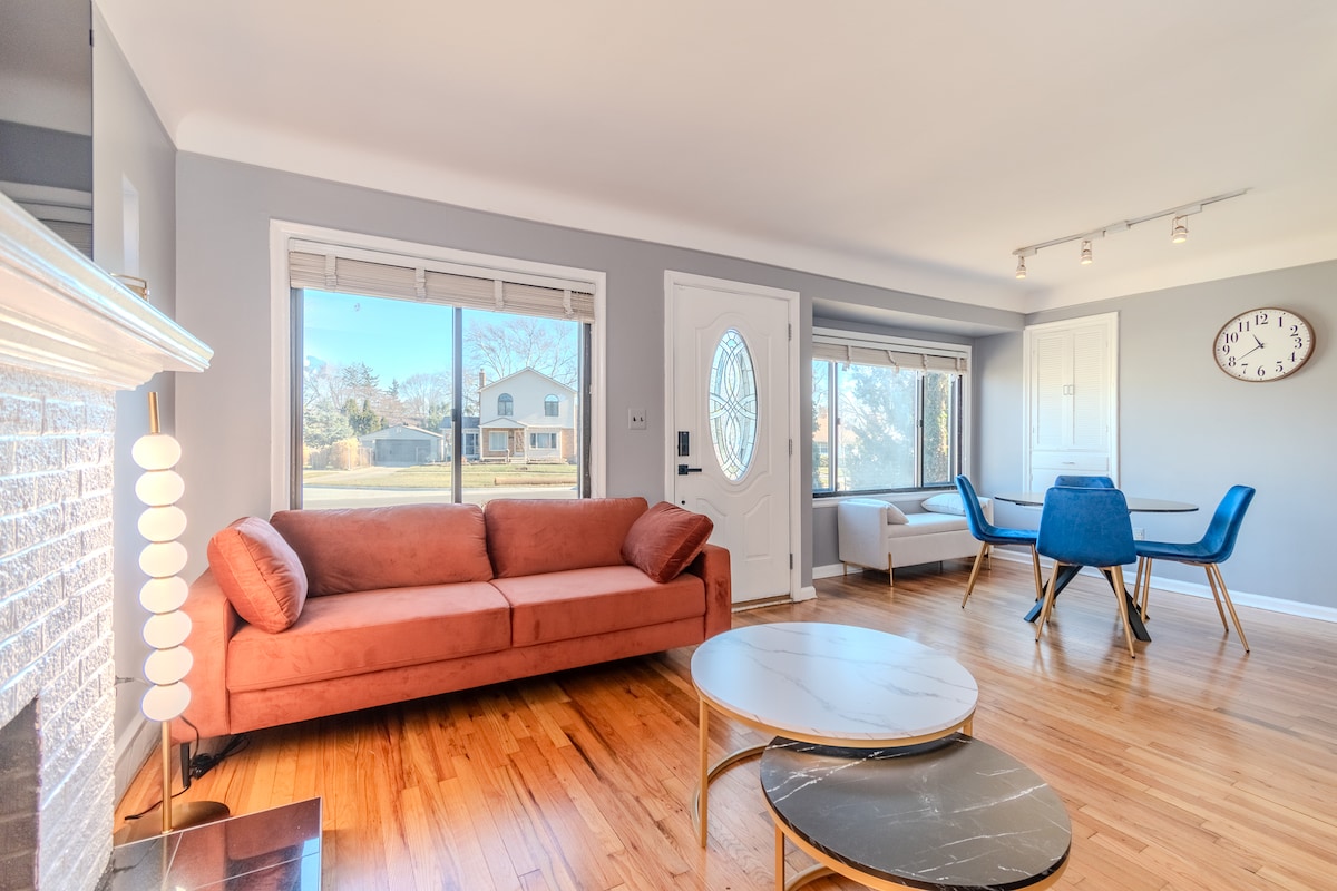 An inviting living area features a plush orange sofa facing a large window that allows natural light to fill the space. A circular coffee table is positioned on the hardwood floor, and a dining area is visible in the background, highlighted by modern blue chairs.