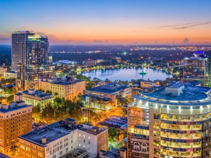 The image captures an aerial view of Orlando at twilight, showcasing a vibrant downtown skyline. Illuminated buildings are reflected in a serene body of water, while the horizon displays soft hues of orange and blue as daylight transitions to night.