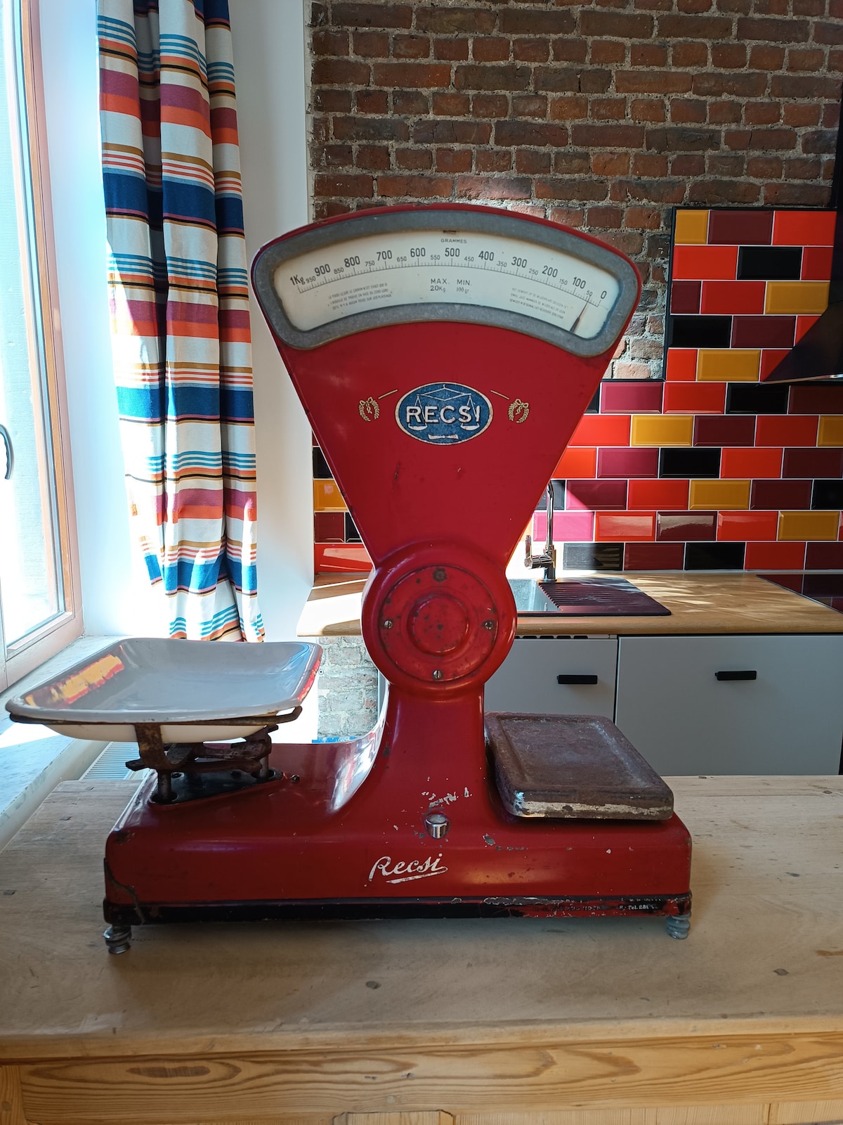 An antique red weighing scale is prominently displayed on a wooden countertop. The scale features a large, distinctive dial and a shallow measuring tray. In the background, colorful tiled walls add a vibrant contrast, and natural light enters through a nearby window.
