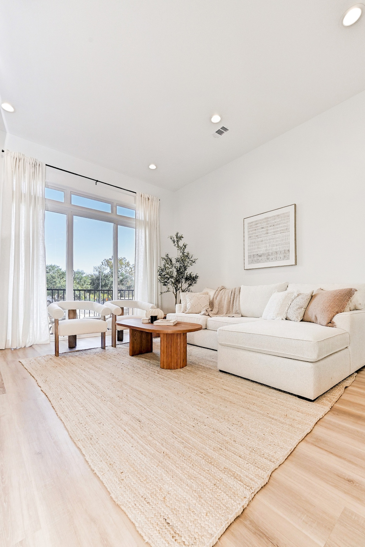 A spacious living area is furnished with a light-colored sectional sofa, accompanied by two armchairs. A natural fiber rug covers the floor, and coffee tables are placed in front. Large windows with sheer curtains allow natural light to fill the room, providing a view of the outdoors.