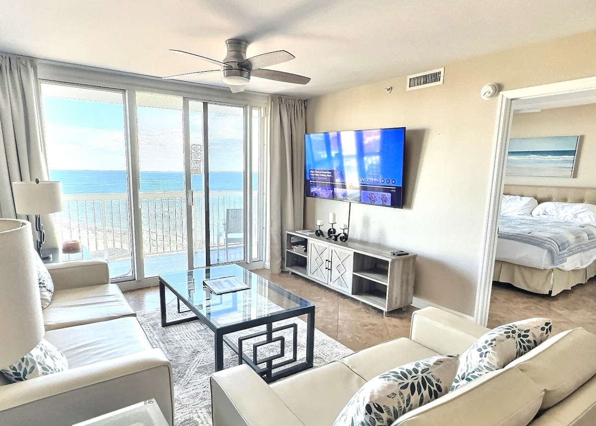 A bright living area features a spacious white leather sofa and a glass coffee table. Floor-to-ceiling windows offer sweeping views of the ocean. A large Smart TV is mounted on the wall, with soft light entering from the balcony. The master bedroom is visible through an open doorway.