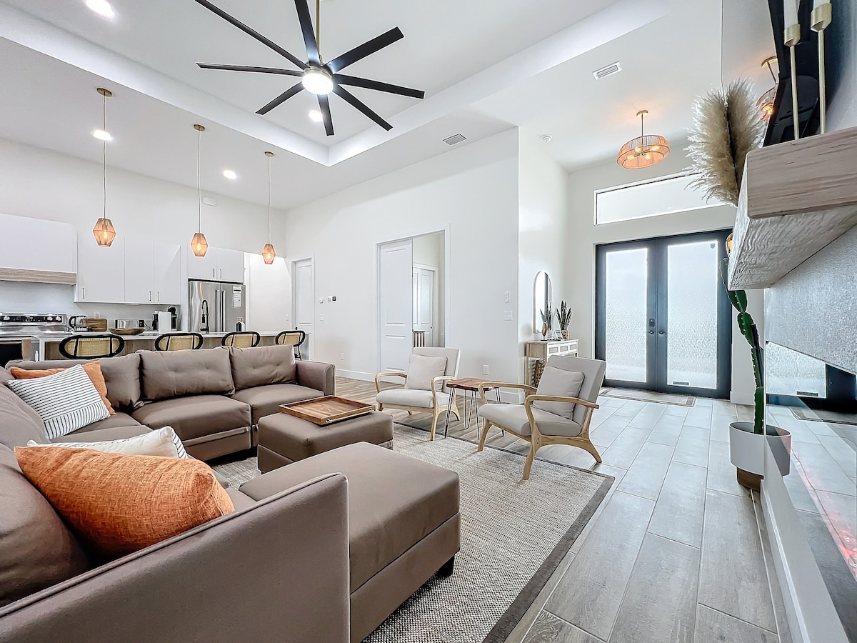 A spacious living room is shown, featuring a sectional sofa complemented by decorative pillows. A large ceiling fan hangs above, and natural light enters through glass doors. Modern pendant lights illuminate the area, which includes a coffee table and two accent chairs, creating an inviting atmosphere.