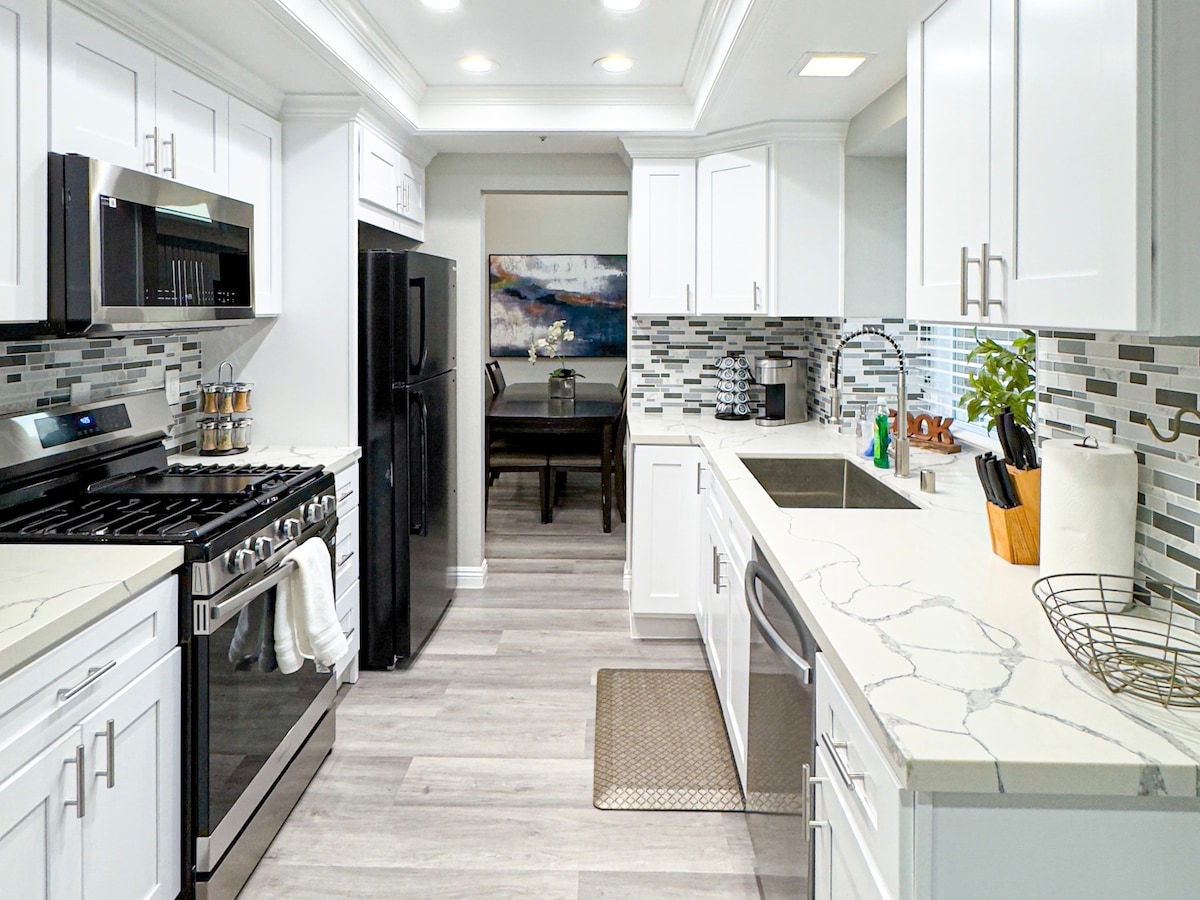 The kitchen features modern appliances, including a gas stove and a double-door refrigerator. Sleek white cabinetry contrasts with the textured backsplash, while stone countertops provide ample workspace. A dining area is visible in the background, enhancing the kitchen's open layout.