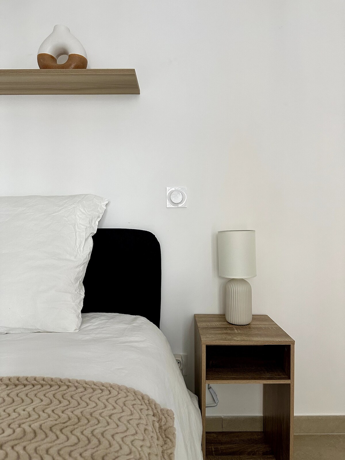 The image displays a cozy sleeping area featuring a spacious bed with white bedding. A light brown throw blanket is casually draped across the bed. A minimalist bedside table holds a textured lamp. A wooden shelf is positioned above the bed, displaying a decorative item.