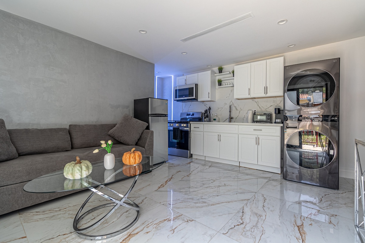 A modern living area showcases a sleek glass coffee table with decorative pumpkins on top. Soft seating is arranged around the table, and a contemporary kitchen is visible in the background, featuring stainless steel appliances and minimalist cabinetry. Light marble flooring complements the neutral color scheme.
