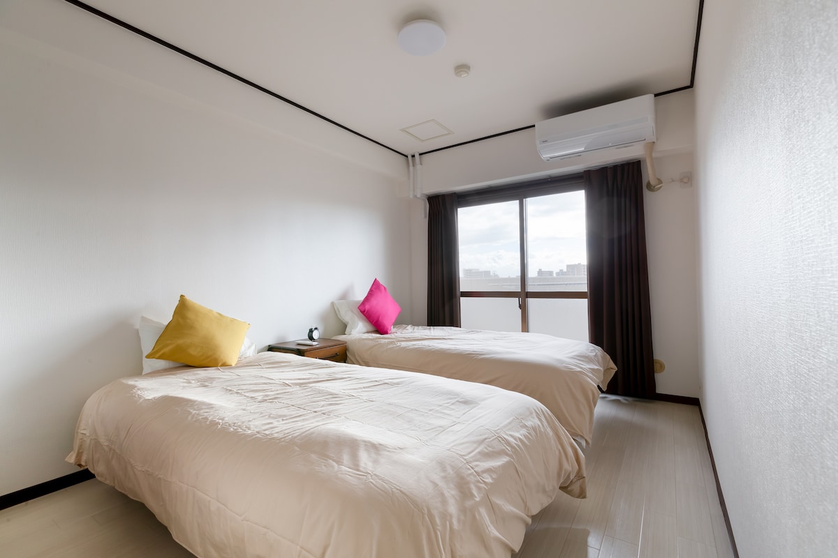 A well-lit guest room features two separate beds dressed in soft beige linens. Decorative cushions in yellow and pink enhance the minimalist decor. Natural light enters through a floor-to-ceiling window framed by dark curtains, and an air conditioning unit is mounted on the wall.