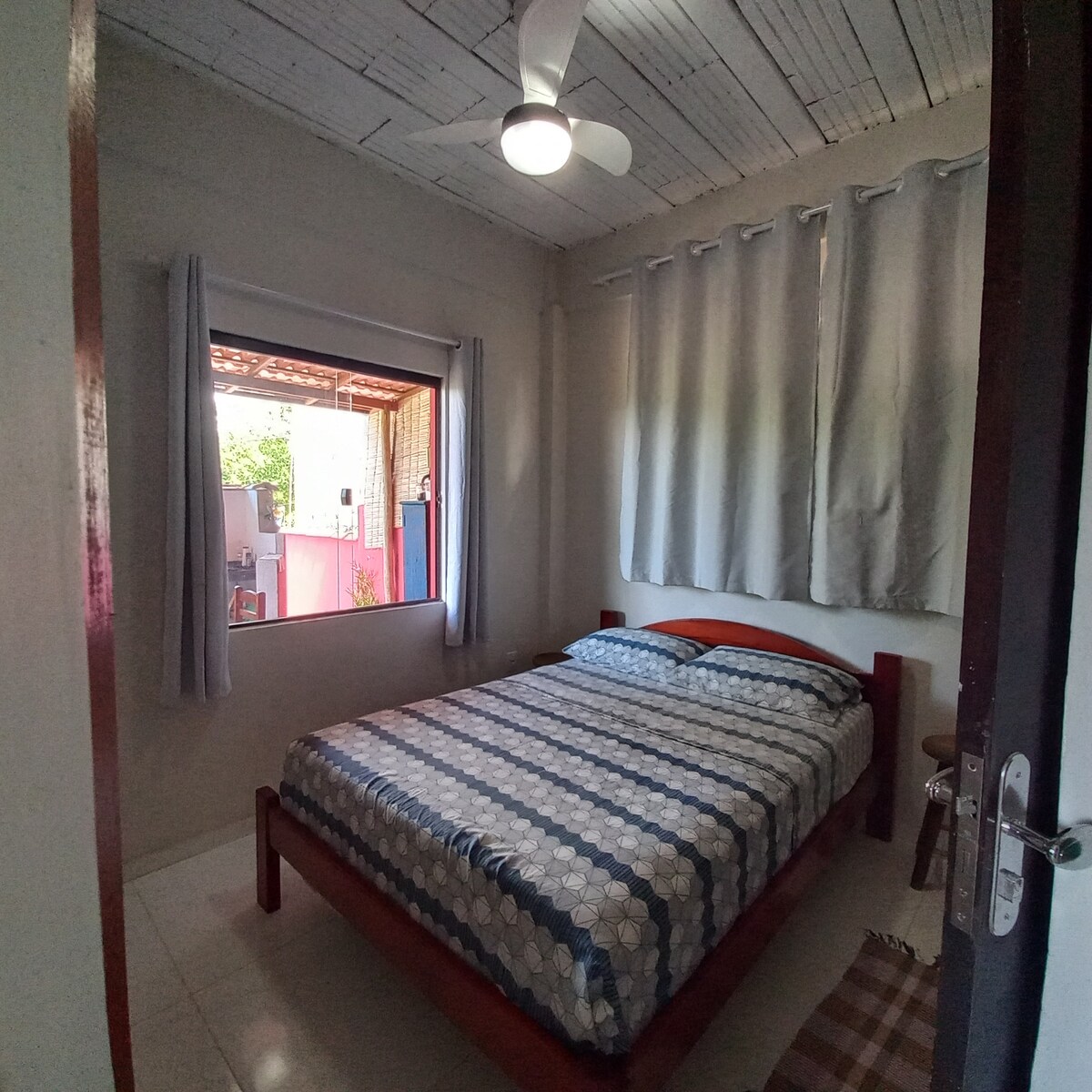 Dormitorio doble de la villa con ventilador de techo, 1 gran ventana de vidrio que conduce al balcón gourmet y 2 vitrums en la pared detrás de la cama. Tenemos un gabinete incorporado con estantes y perchas que no aparecían en la foto.
