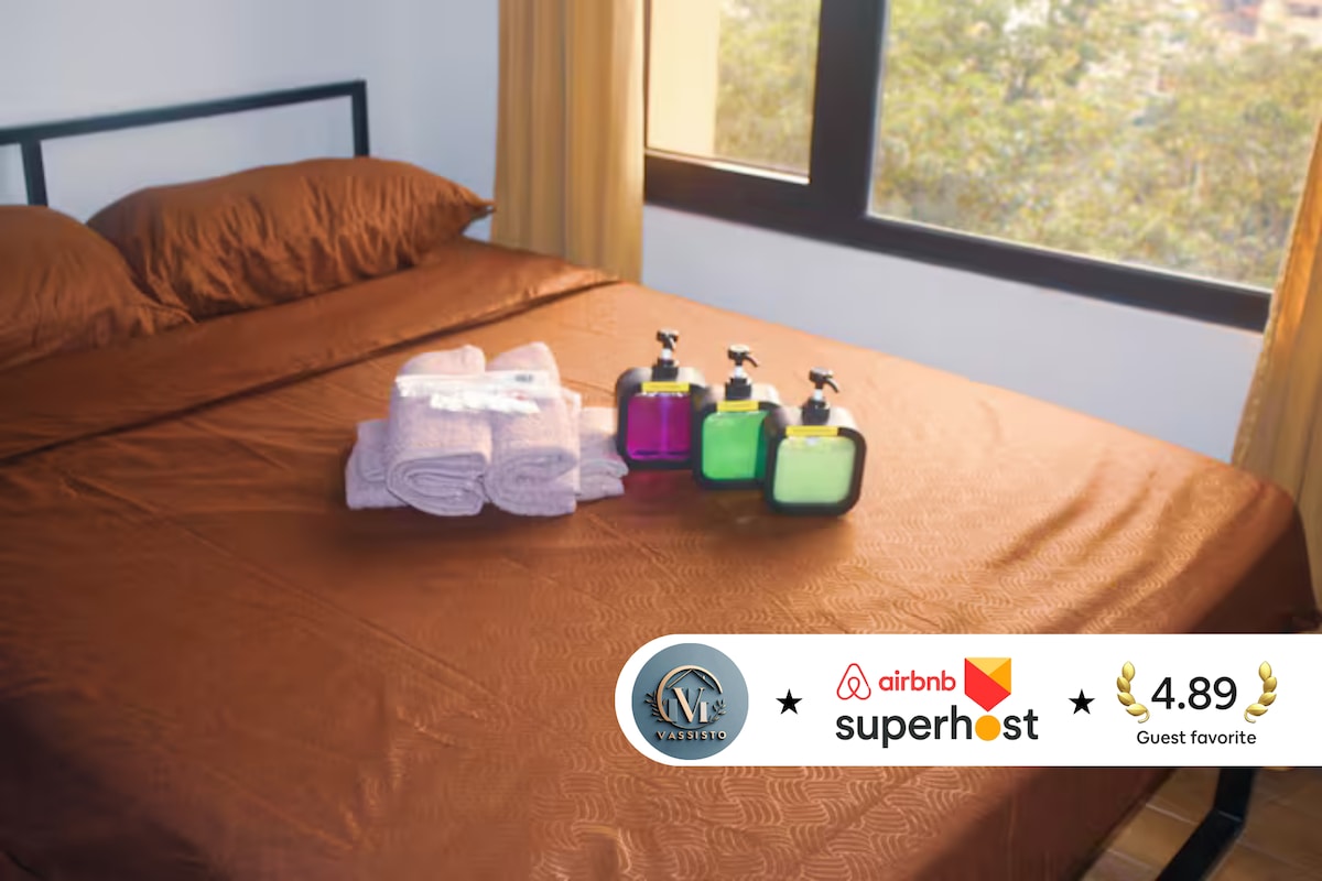A spacious bed is dressed in bronze-colored linens, complemented by two matching pillows. Neatly arranged on the bed are pairs of pastel bath towels and three dispensers of toiletries, featuring vibrant colors. Natural light filters through the nearby window, enhancing the warmth of the space.