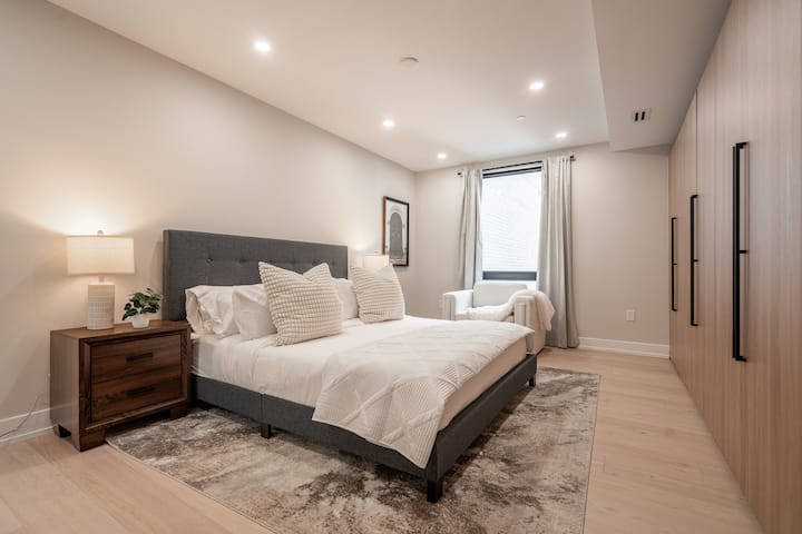 Pokojná spálňa s jemnými neutrálnymi tónmi s pohodlnou posteľou s plyšovými vankúšmi, drevenými nočnými stolíkmi a útulným kreslom pri okne.