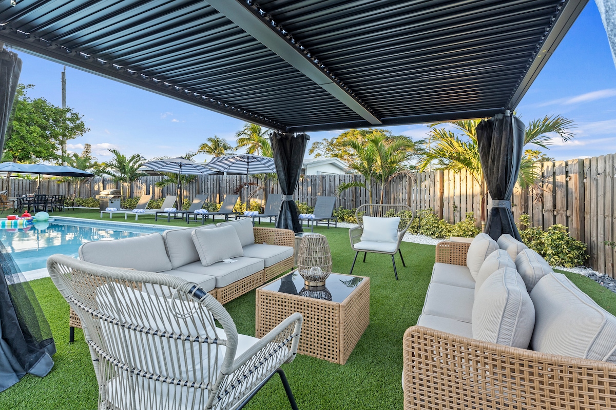 An outdoor seating area is situated under a retractable pergola, featuring a blend of woven and plush chairs surrounding a glass-top coffee table. The space overlooks a large pool and is framed by palm trees and umbrellas, creating a relaxed atmosphere for gatherings.