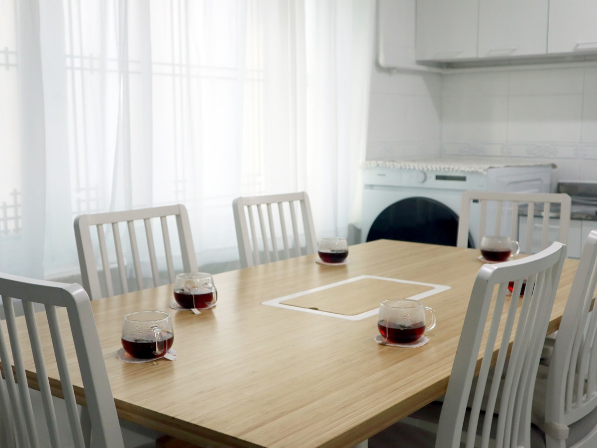 A spacious dining table is set in a well-lit area, featuring six white chairs. The table is topped with glasses filled with a dark beverage. Soft white curtains allow natural light to filter in, creating a bright and welcoming atmosphere.