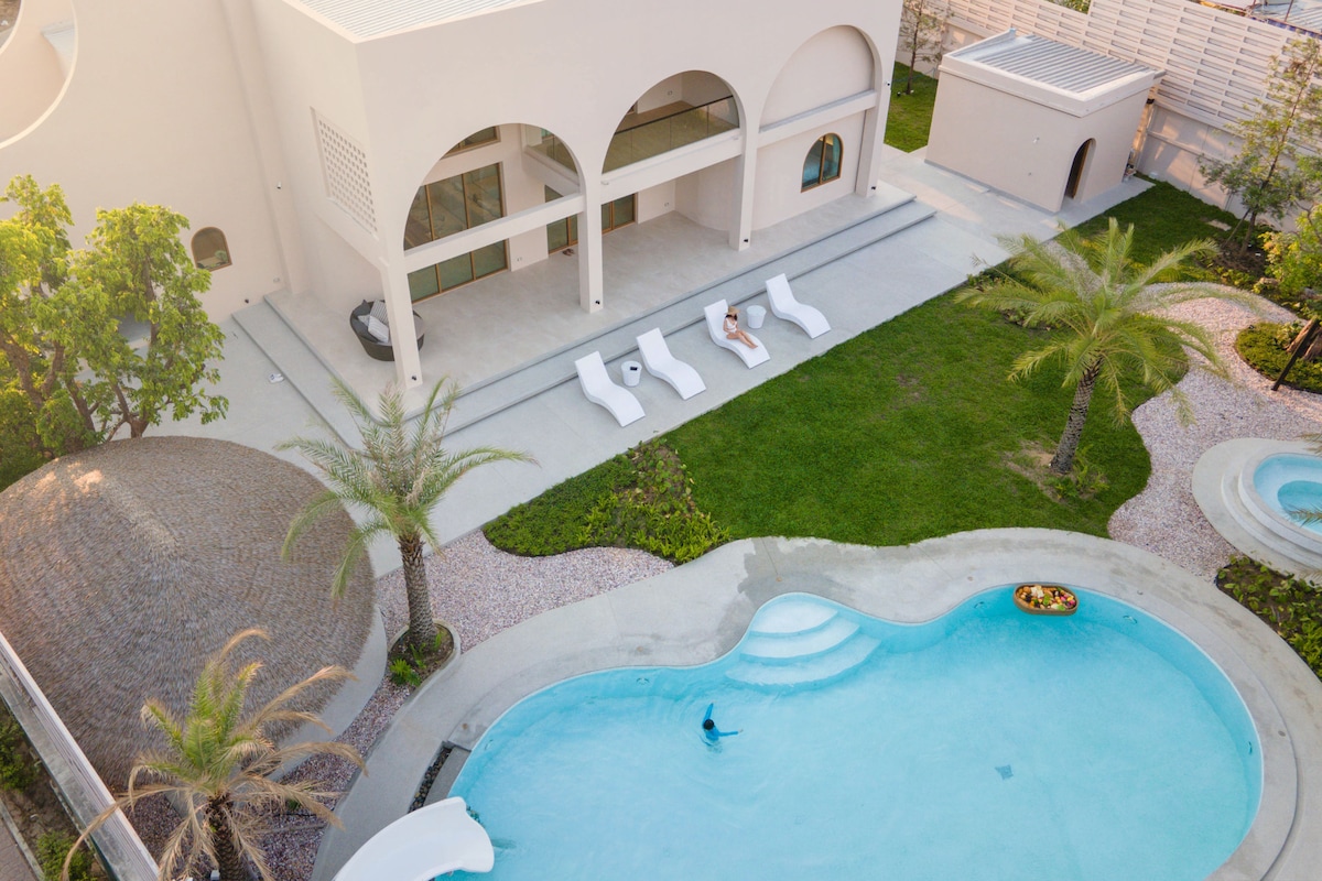 An aerial view showcases a spacious outdoor area featuring a large private pool with a curved shape. White loungers are positioned around the pool, surrounded by green landscaping and palm trees. A shaded seating area is visible, contributing to the serene atmosphere.
