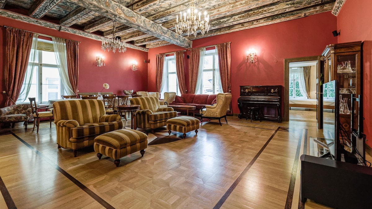 A spacious living and dining area features rich red walls and exposed wooden beams. Elegant chandeliers illuminate the room, which contains plush seating including striped armchairs and a cozy sofa. Large windows provide ample natural light, enhancing the space's inviting atmosphere.