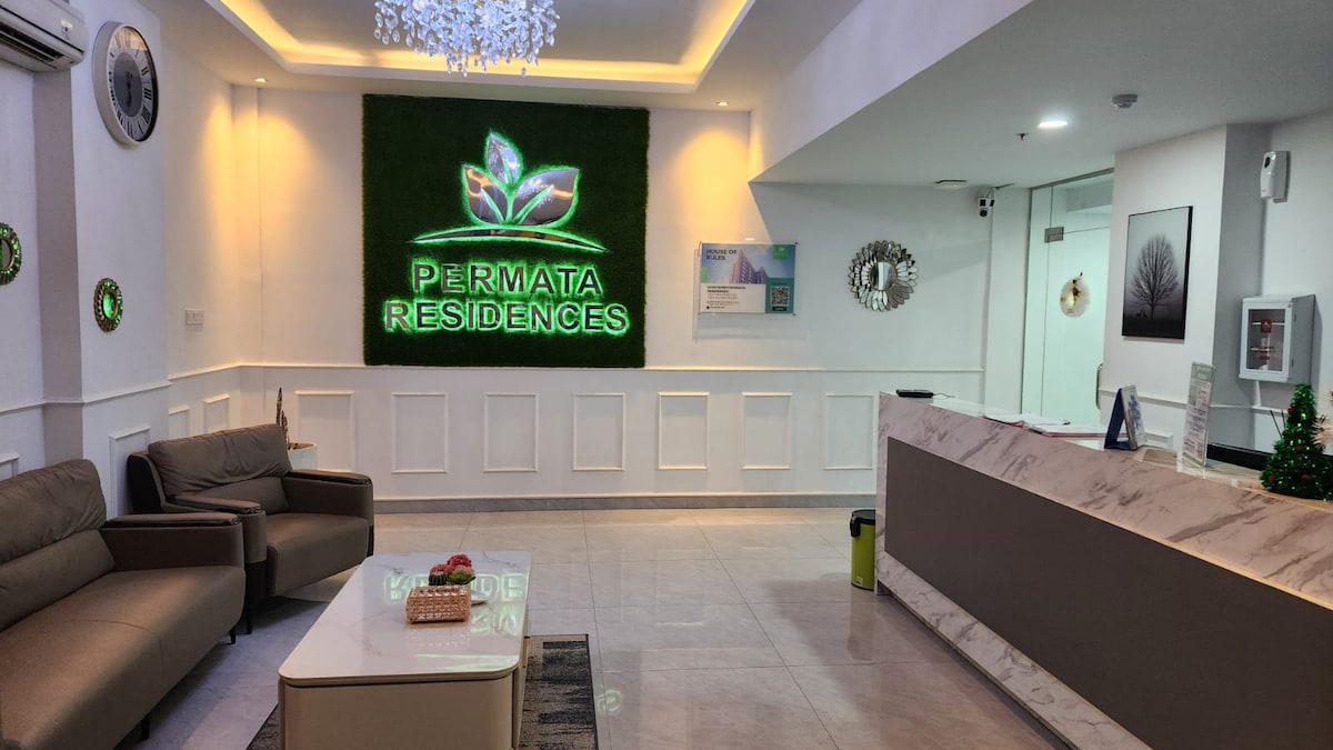 The lobby area features a modern reception desk with a marble finish. Comfortable seating arrangements are provided on either side, with a prominent green wall displaying the logo 'Permata Residences' illuminated above. Soft lighting creates a welcoming ambiance.