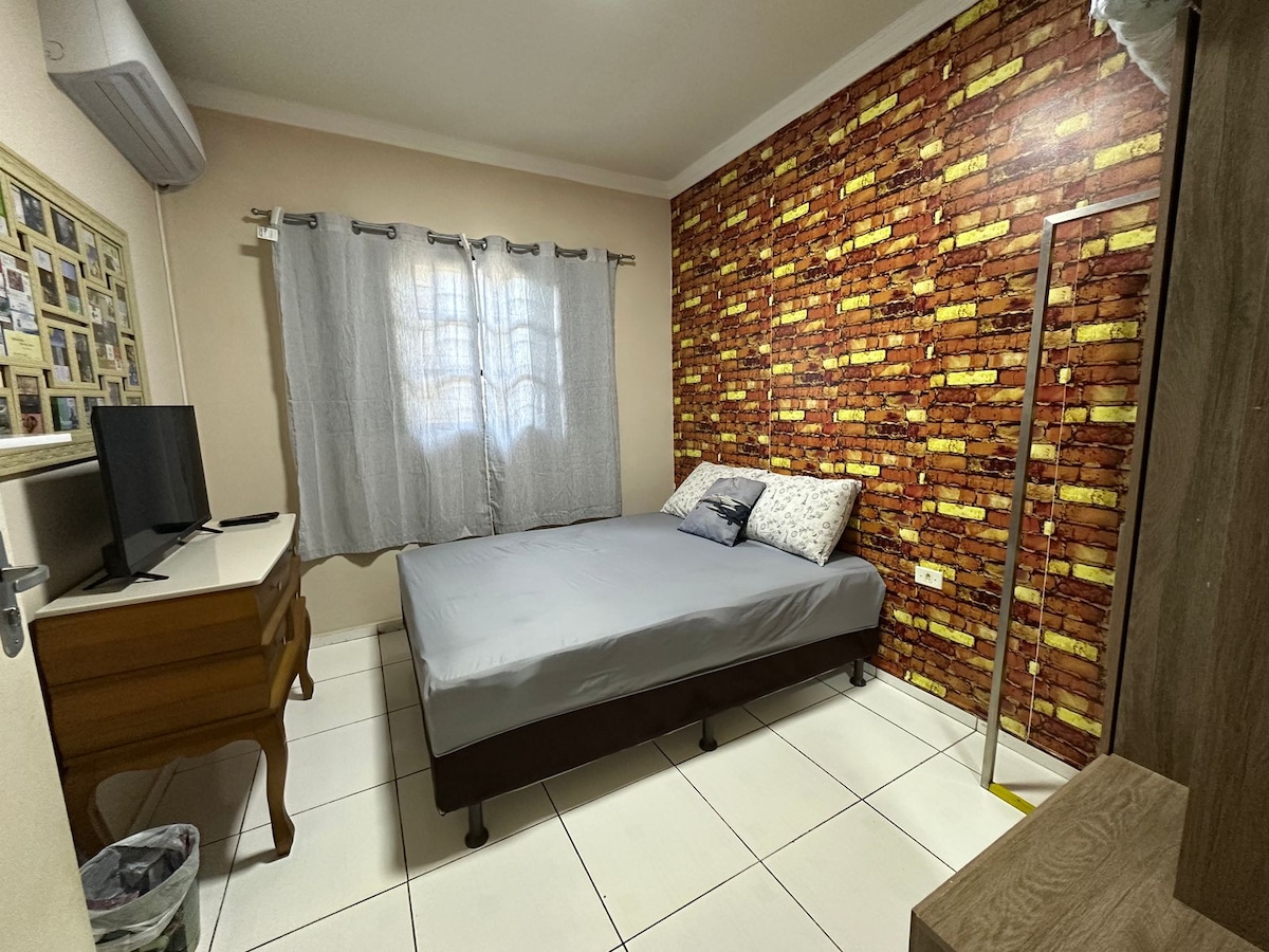 A well-furnished bedroom features a spacious bed dressed in neutral linens. The room is enhanced by a feature wall with a brick pattern and soft curtains covering a window. A wooden dresser and a small television are included, providing essential comforts.