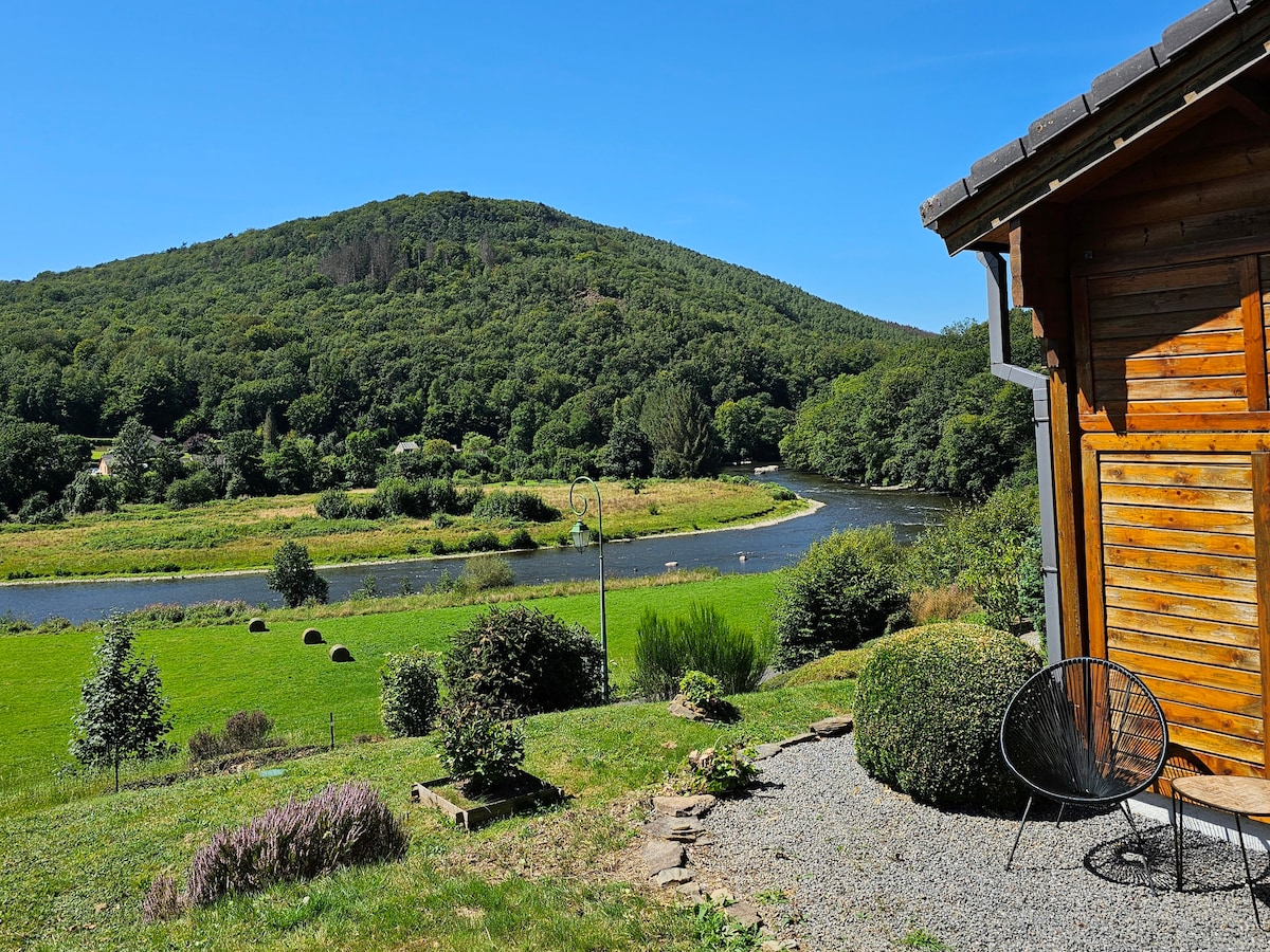 The image displays a scenic view from a wooden chalet, featuring a lush green landscape with rolling hills and a gently curving river. The foreground includes gravel and shrubs, while the backdrop showcases vibrant trees under a clear blue sky.