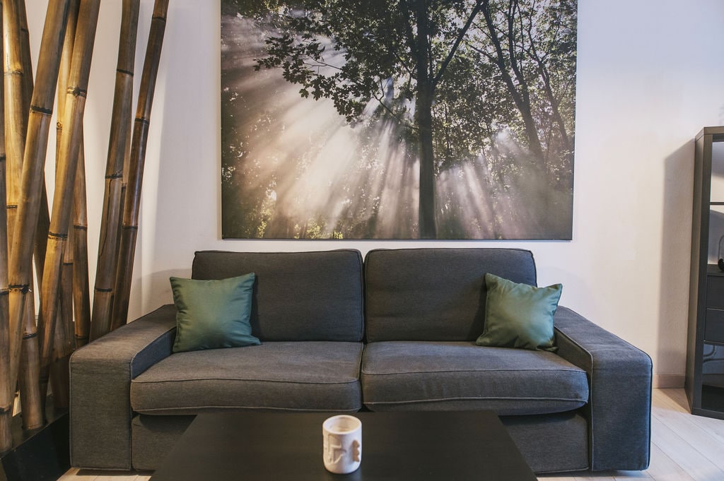 A modern living area features a dark gray sofa adorned with green decorative pillows. A coffee mug is placed on a simple black coffee table. Behind the sofa, a large artwork depicting trees with rays of sunlight provides a serene backdrop.