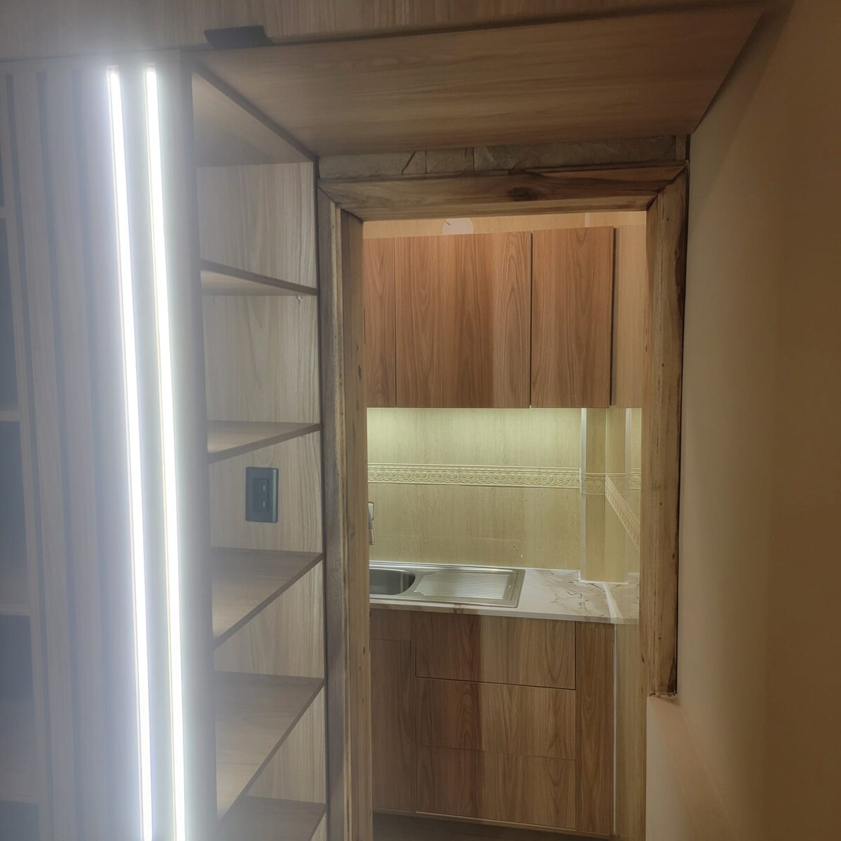 A modern kitchen is visible through an open doorway, featuring sleek wooden cabinetry and a polished countertop. Soft lighting creates a warm glow, illuminating the space where a stainless steel sink sits under wall-mounted cabinets. Shelving beside the entrance is lined with light strips.
