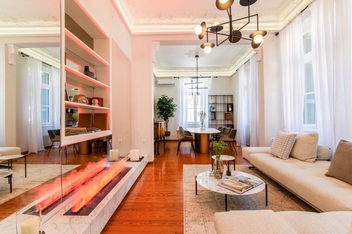 A modern living area is highlighted by a stone fireplace with a vibrant flame effect. Contemporary furniture, including a light-colored sectional sofa and a marble coffee table, complements the space. Natural light pours in through large windows, enhancing the open ambiance and stylish décor.