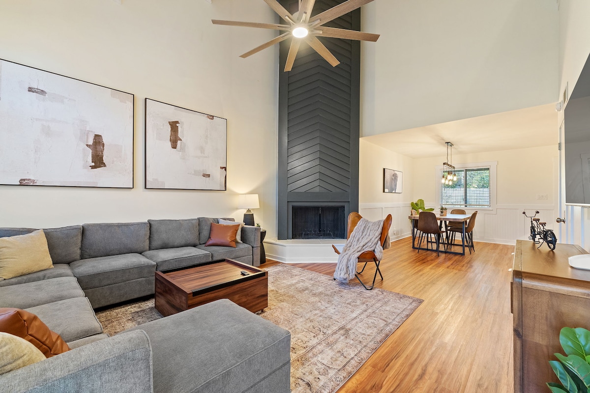 A spacious living room features a sectional sofa and a wooden coffee table. Above, a large ceiling fan rotates. Natural light filters through a nearby window, illuminating the artwork on the walls. An adjacent dining area is visible, showcasing a wooden table and chairs.