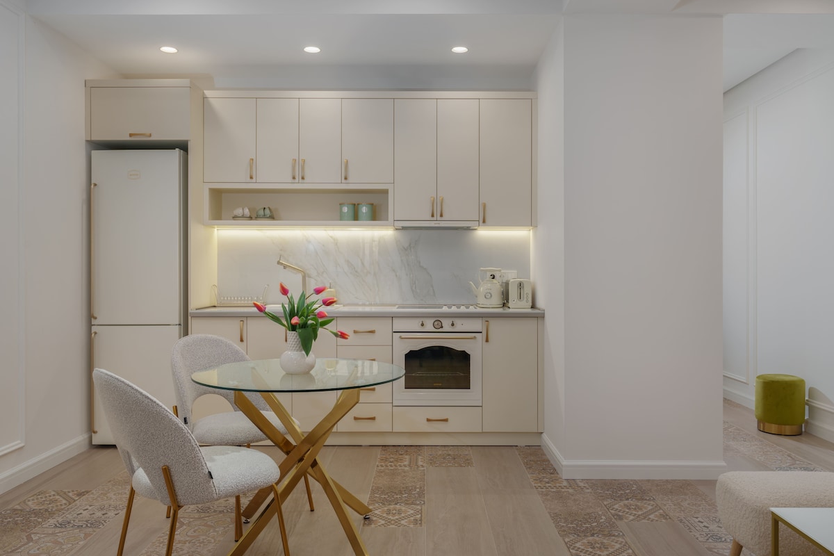The kitchen area features light cabinetry with marble accents and modern appliances, including a refrigerator and oven. A glass dining table is surrounded by two upholstered chairs. Soft lighting enhances the clean, contemporary design, offering a welcoming space for meals or gatherings.