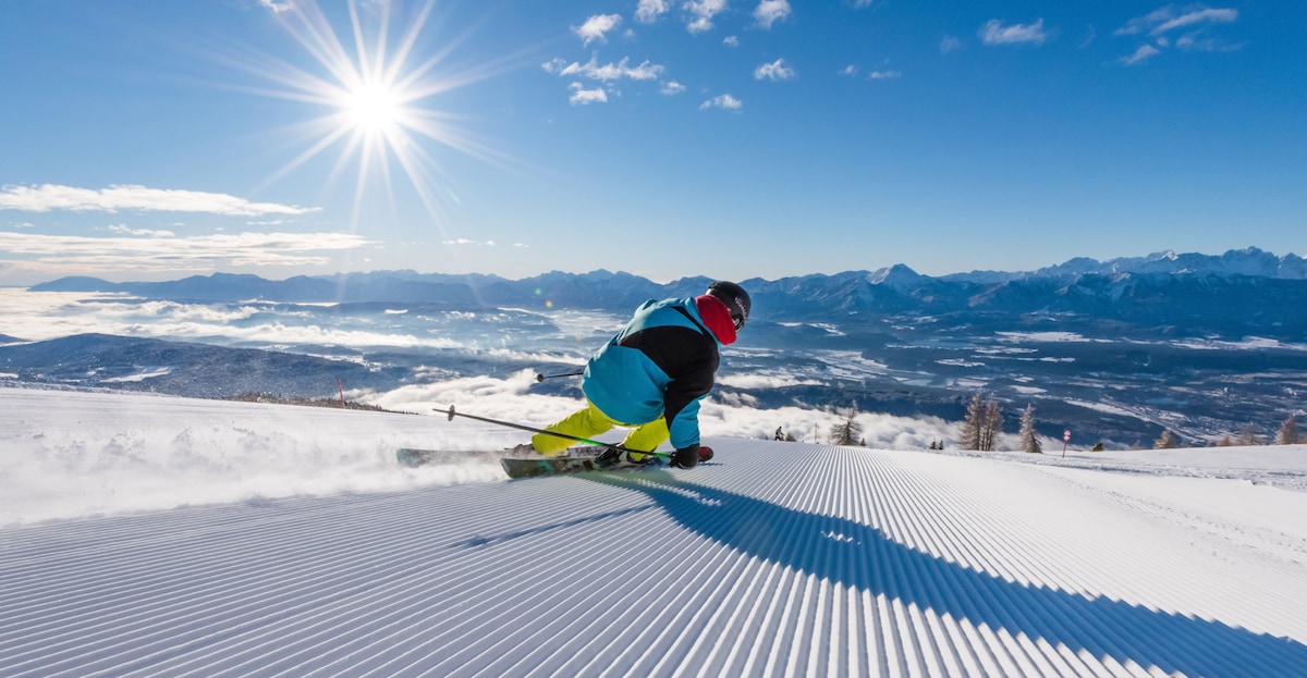 A snowboarder glides down a groomed snow slope under a bright sun. The background reveals mountains and valley fog, with a clear blue sky above. The snow-covered landscape reflects the sunlight, creating a serene winter atmosphere.