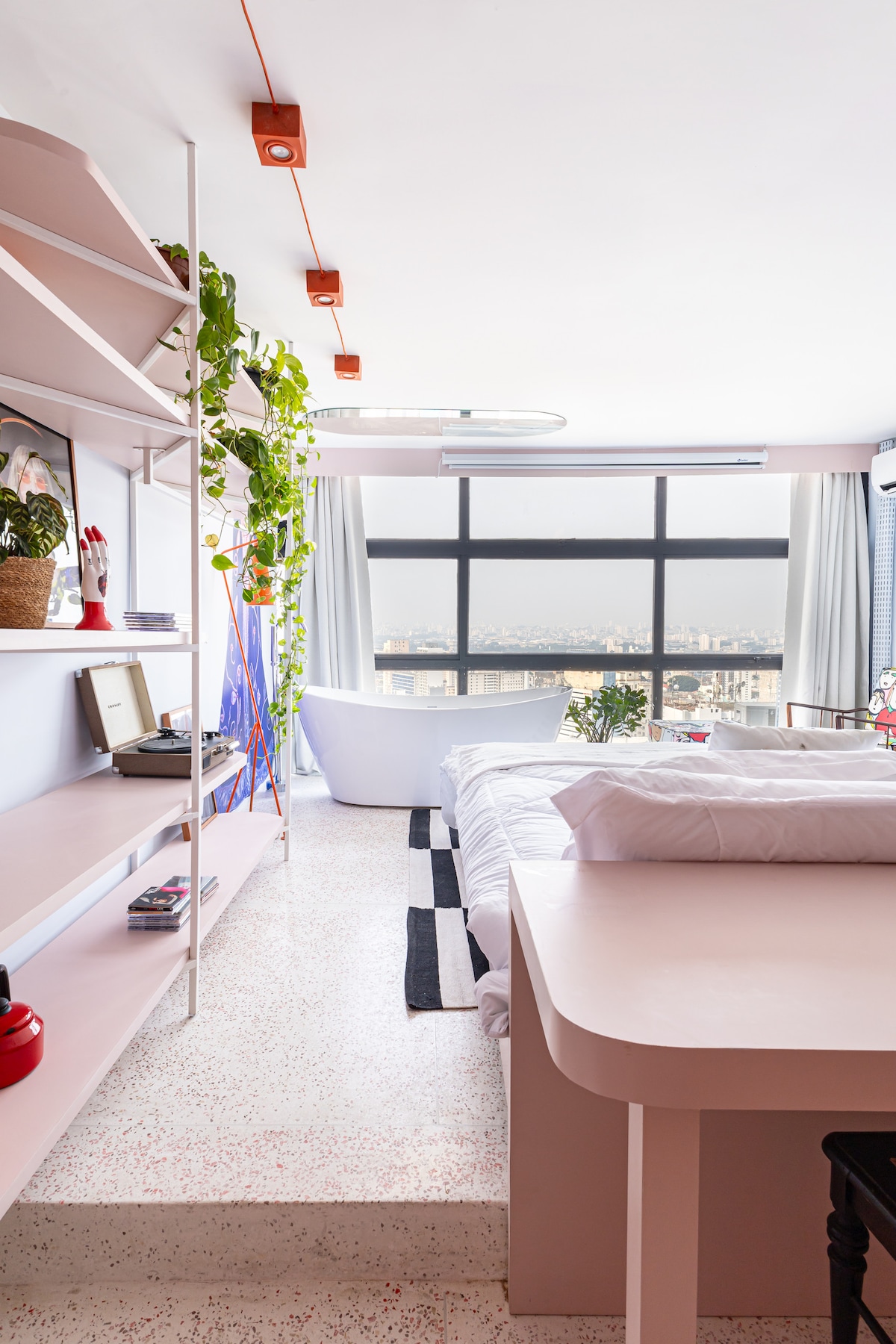 A bright studio space features a spacious bed with crisp white linens, complemented by decorative plants and shelves holding vintage items. Large windows provide panoramic city views, while sunlight fills the room, enhancing the light-colored décor and modern furnishings.