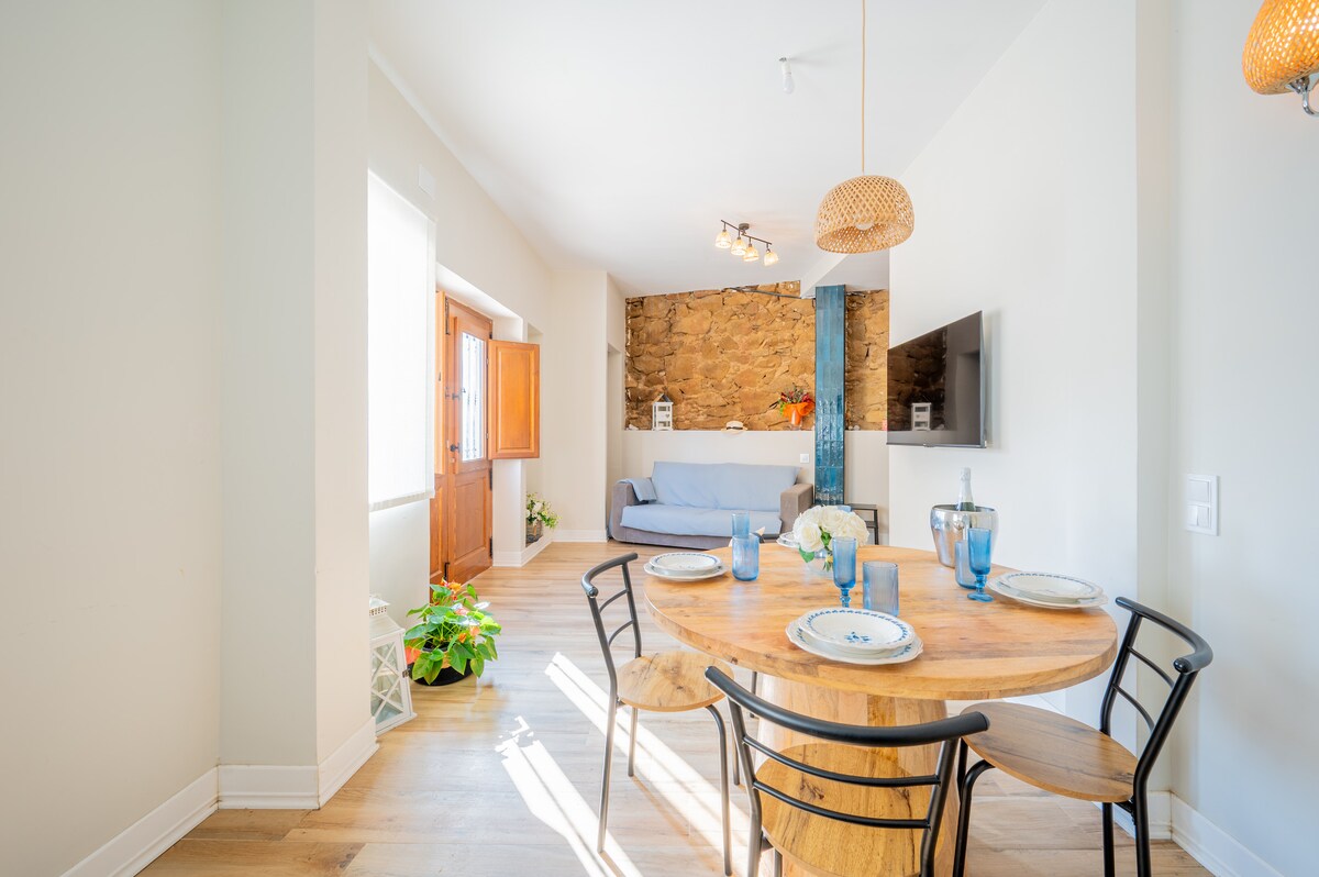 An inviting dining area features a round wooden table set with plates and blue glasses. A comfortable sofa in the background is complemented by exposed stone walls, creating a cozy atmosphere. Natural light from large windows brightens the space.