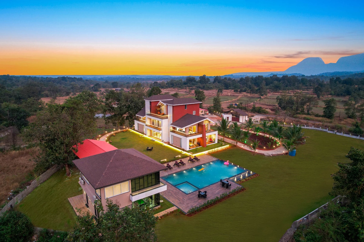 The Grand Crest showcases its impressive facade against a twilight sky, surrounded by lush green lawns. The outdoor pool is gently illuminated, reflecting the serene ambiance. Multiple outdoor seating areas invite relaxation, while the distant hills form a tranquil backdrop.