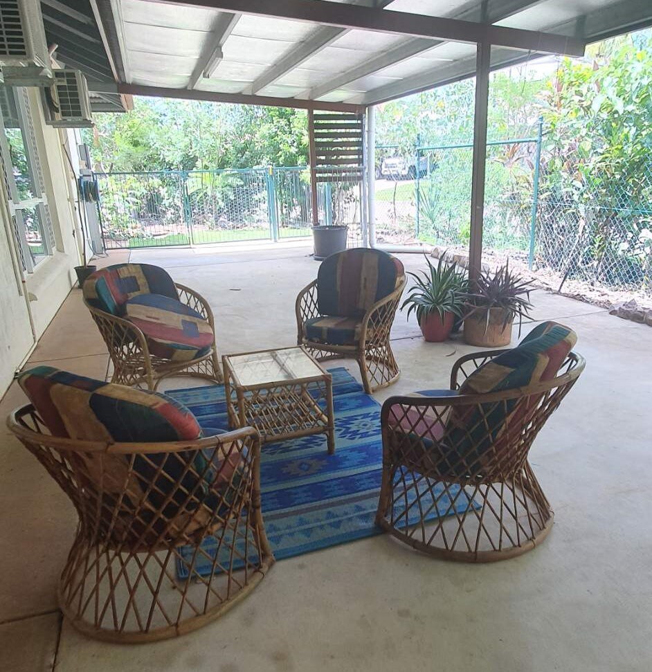 A covered outdoor seating area is showcased, featuring four rattan chairs with colorful cushions arranged around a glass-topped table. A patterned blue rug enhances the space, while greenery from nearby plants adds a refreshing touch to the atmosphere.