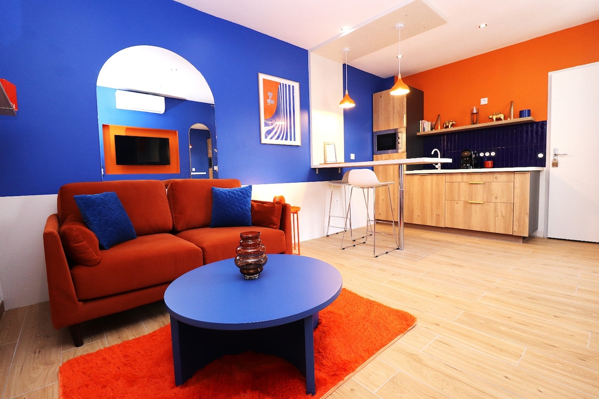 A cozy living area features a vibrant blue and orange color scheme. A spacious orange sofa with blue pillows faces a round blue coffee table, resting on a plush orange rug. In the background, a modern kitchen with wood cabinetry and a breakfast bar is visible.