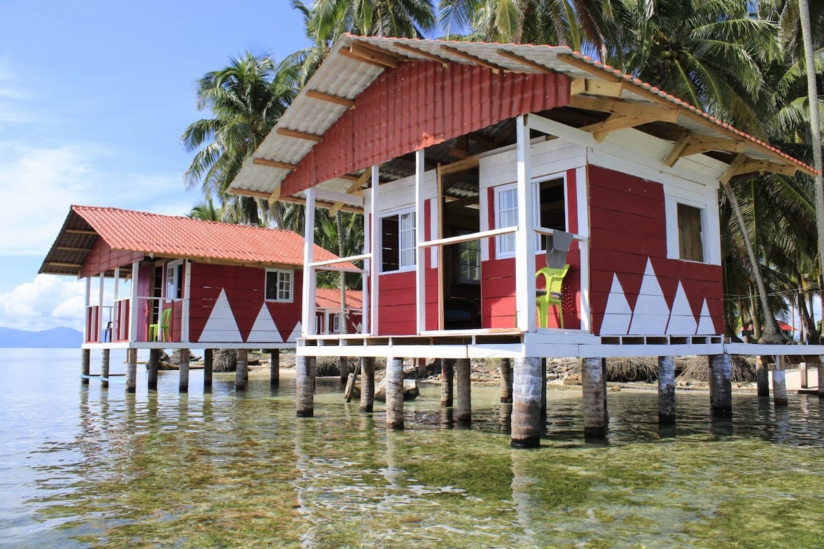Two overwater cabins stand on stilts above clear, shallow water, featuring red and white wooden exteriors. Each cabin has a porch with vibrant green chairs, surrounded by lush palm trees and the tranquil Caribbean backdrop. The serene atmosphere is enhanced by the gentle rippling of water.