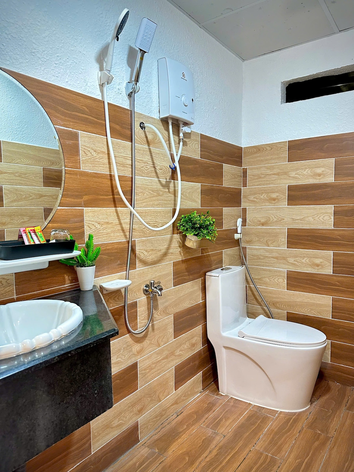A modern bathroom features a combination of wooden and neutral tiles, creating a clean and functional space. A round mirror is mounted above a sleek sink, while a shower with a handheld nozzle is positioned next to a compact toilet. A small plant adds a touch of greenery.