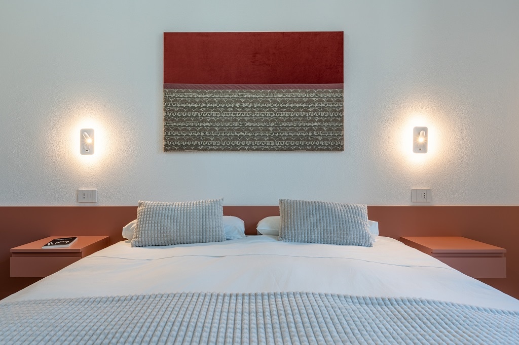 A bright sleeping area features a spacious bed with two textured pillows. The headboard is adorned with a contemporary piece of art in varying shades of red and beige. Soft lighting emanates from two wall sconces, enhancing the minimalist design.