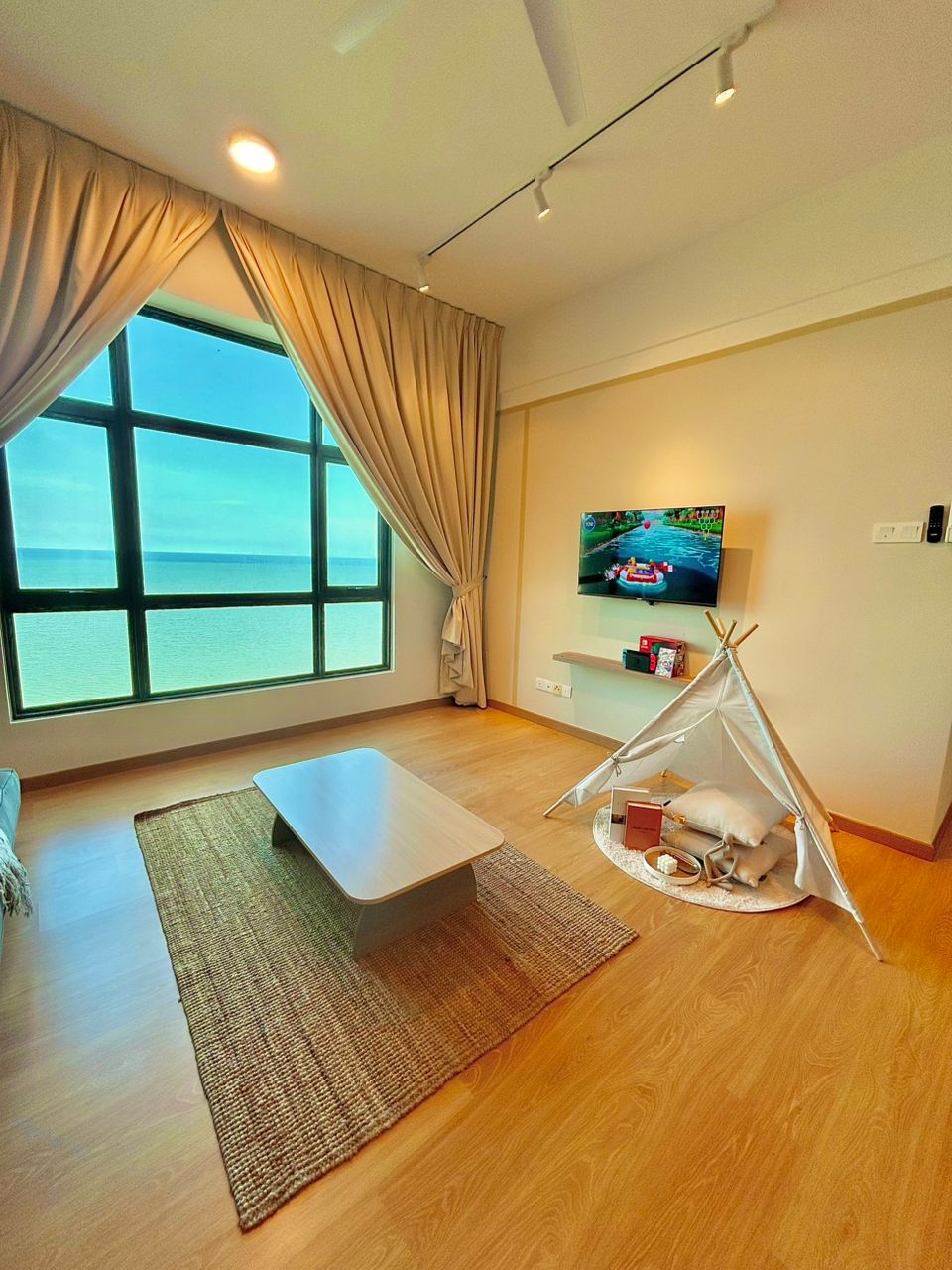 A bright living area features large windows allowing natural light to flood in. A flat-screen television is mounted on the wall, with a small coffee table placed on a neutral rug. A cozy play tent is tucked into one corner, adding a playful touch.