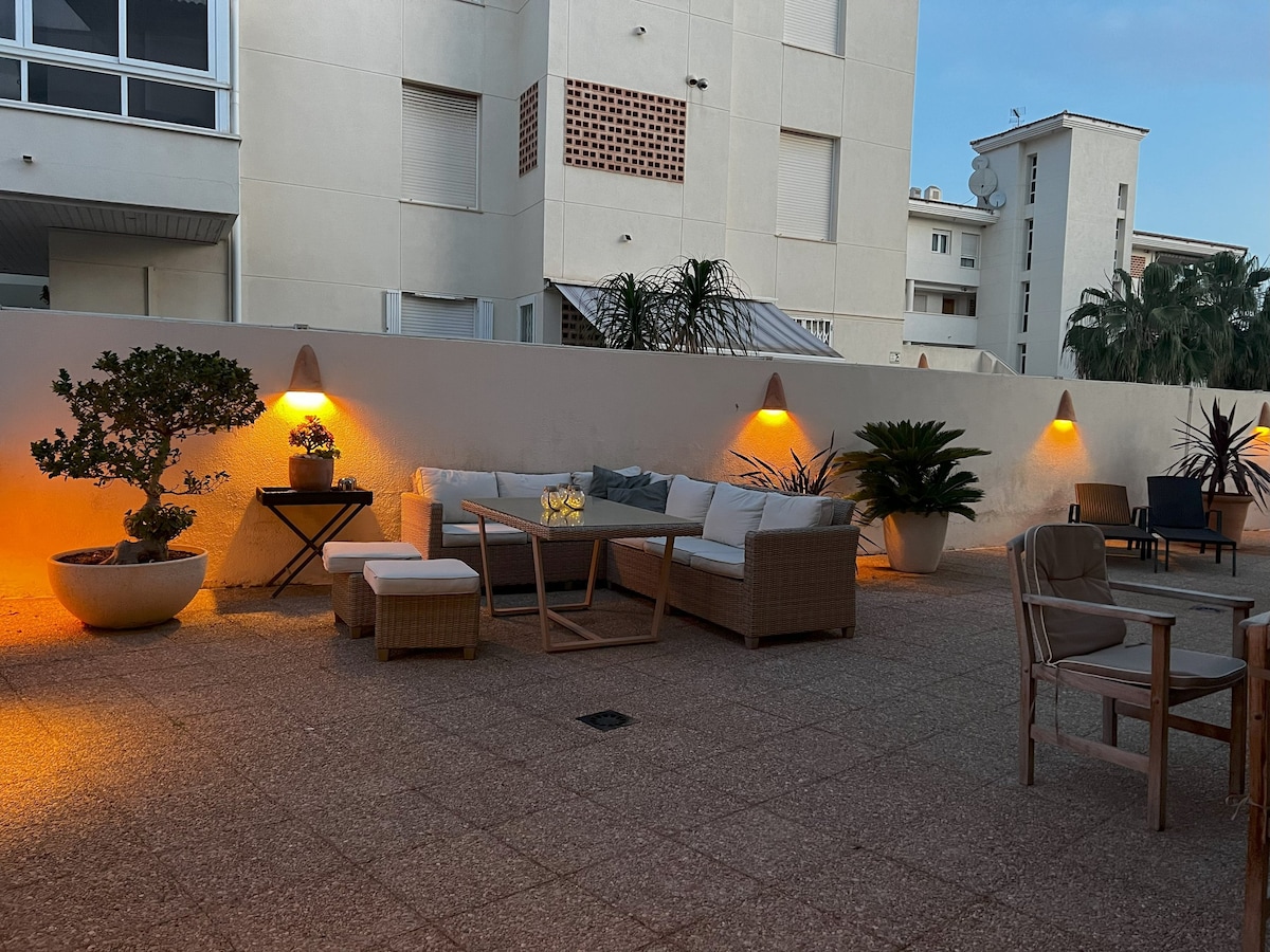 A spacious terrace area is displayed, featuring comfortable seating arrangements made from woven materials. Soft lighting from wall-mounted fixtures creates a calming ambiance. Potted plants add greenery, enhancing the outdoor setting. The surface is paved, providing a clean and accessible space for relaxation.