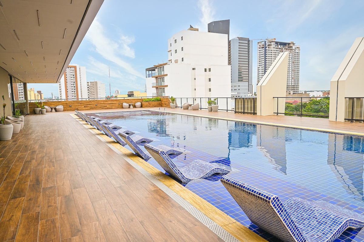 An expansive rooftop pool area is depicted, featuring lounge chairs positioned along the water's edge. The pool's blue tiles mirror the clear sky, while the surrounding city skyline provides a modern backdrop. Potted plants line the space, adding a touch of greenery.