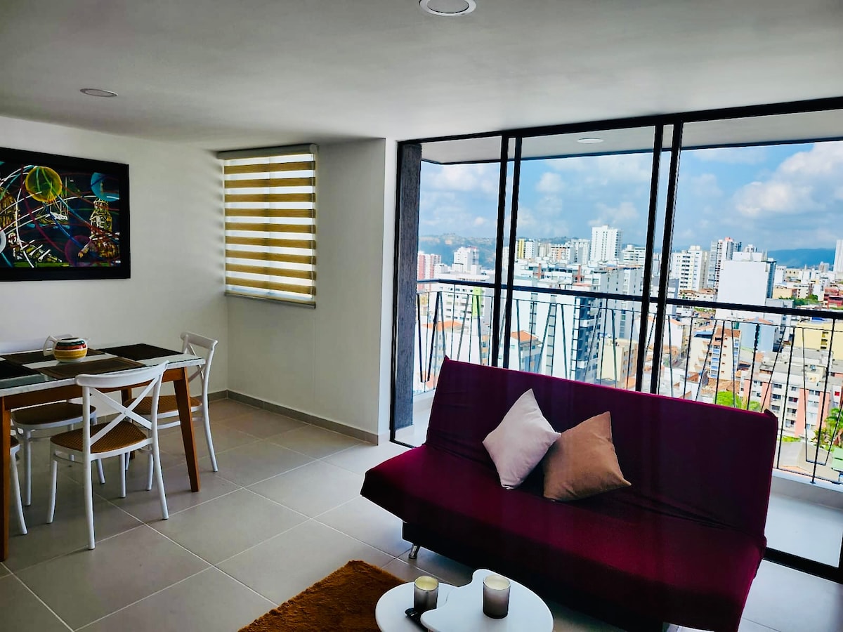 A modern living space features a burgundy sofa with decorative pillows, a dining area with a table and chairs, and large windows providing city views. Light filters in through the partially closed blinds, highlighting the textured wall art and cozy atmosphere.