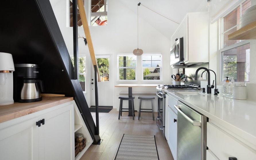 A modern kitchen is featured with sleek white cabinetry and stainless steel appliances. A wooden countertop and coffee maker are positioned near a window, while a small dining table with two stools is situated beside it, allowing natural light to fill the space.
