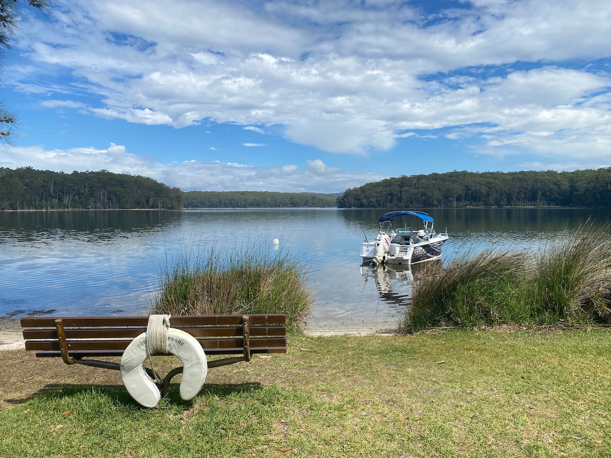 A serene lake view is displayed, featuring a grassy shore with a wooden bench in the foreground. A white life ring is attached to the bench. In the background, a boat is moored by the tranquil water, surrounded by lush greenery and under a partially cloudy sky.