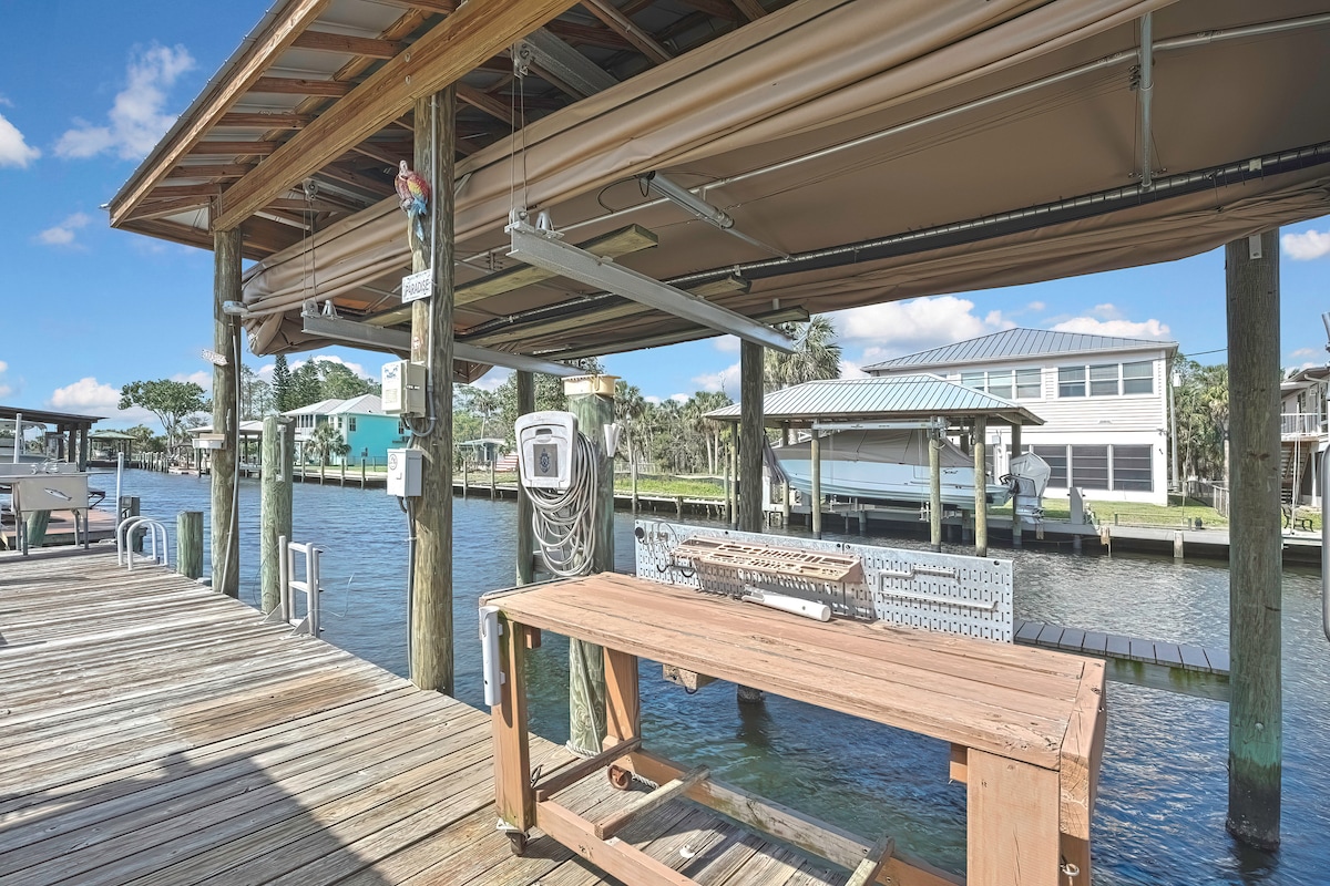 A wooden dock sits along the water's edge, featuring a shaded area with overhead covering. Boat utilities are visible, including a power source and cleaning station. The calm water reflects the surroundings, with nearby homes and lush greenery providing a scenic backdrop.