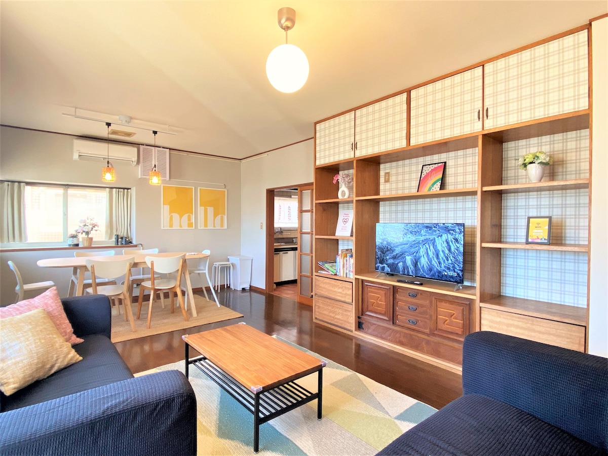 The living area displays a comfortable layout, featuring two dark sofas and a coffee table on a patterned rug. A wooden cabinet holds a television, while warm lighting hangs above the dining table set for six. Natural light enters through a window, complementing the decor.